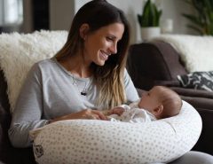 A new mom smiling while breastfeeding her baby on a curved memory foam nursing pillow, showcasing ergonomic support in a cozy living room setting.