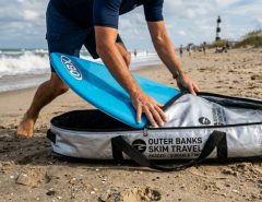 Person packing a blue skimboard into a padded silver travel bag on North Carolina sand.