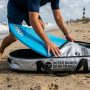 Person packing a blue skimboard into a padded silver travel bag on North Carolina sand.